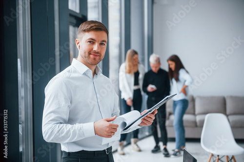 Man in formal clothes standing with documents in front of young successful team that working and communicating together indoors in office