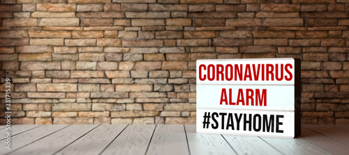 Plakat lightbox with text CORONAVIRUS ALARM #STAYHOME in front of a brick wall on wooden floor