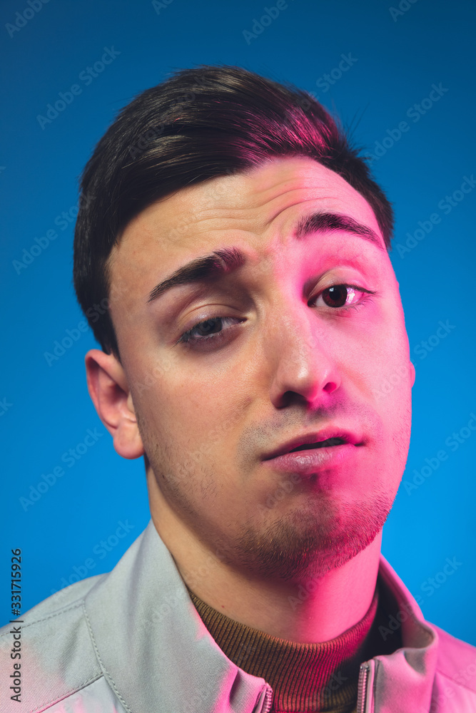 Fototapeta premium Gremaces, looking on. Close up caucasian man's portrait isolated on blue studio background in pink neon light. Beautiful male model. Concept of human emotions, facial expression, sales, ad. Copyspace.
