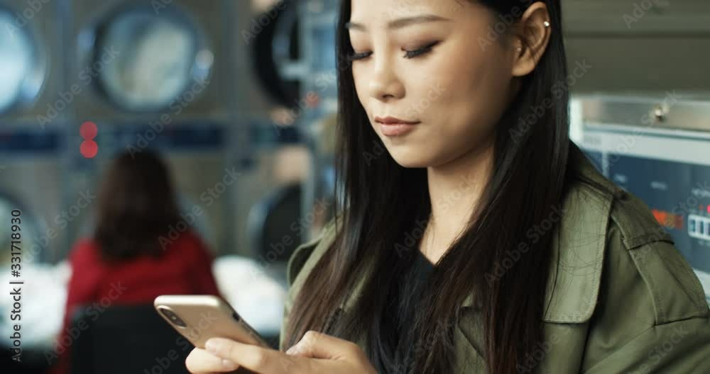 Close up of Asian pretty woman with long dark hair tapping and texting message on smarphone while standing in laundry service room. Beautiful woman typing on phone and waiting for clothes to wash. Close up of Asian pretty woman with long dark hair tapping and texting message on smarphone while standing in laundry service room. Beautiful woman typing on phone and waiting for clothes to wash.