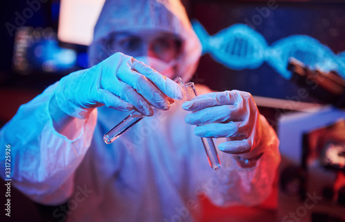 Nurse in mask and white uniform, holding tube with liquid and sitting in neon lighted laboratory with computer and medical equipment searching for Coronavirus vaccine photo