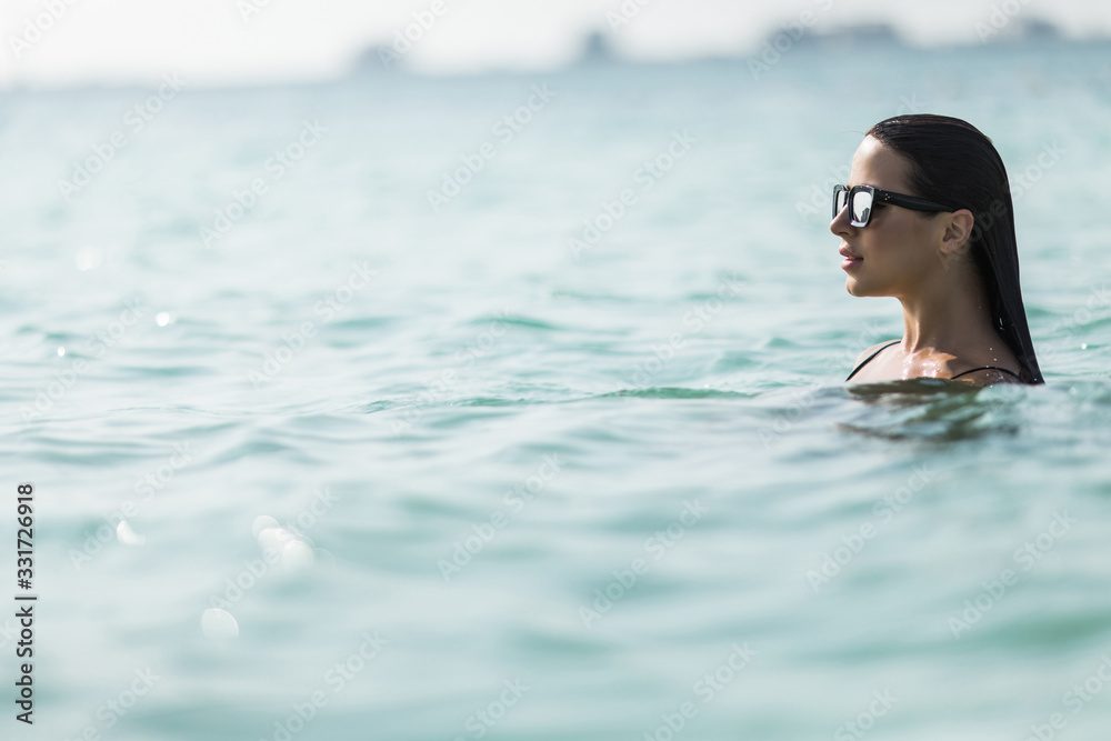 Stunning fit girl in sunglasses posing in sea water and enjoying sun during summer holidays on the tropical beach. Summer lifestyle portrait.