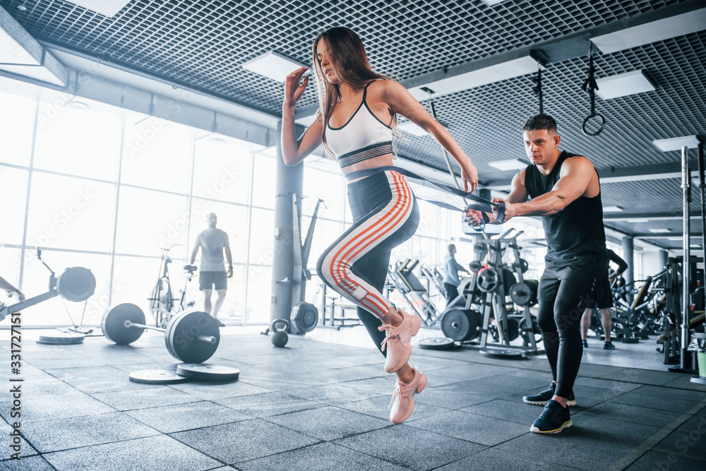 Young sportive people doing exercises together in the gym. Woman running when man holding her by elastic tape Young sportive people doing exercises together in the gym. Woman running when man holding her by elastic tape