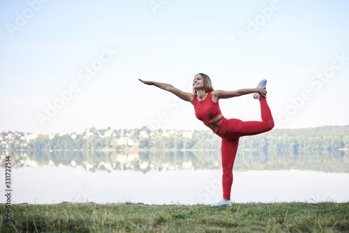 Young blond woman, wearing red leggings, top and white sneakers, doing morning exercises by lake in summer. Fitness yoga stretching training outside by water. Healthy life concept. Active way of life