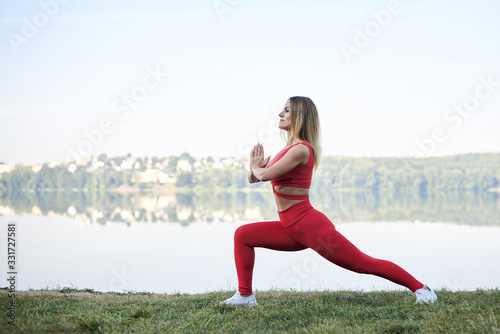 Young blond woman  wearing red leggings  top and white sneakers  doing morning exercises by lake in summer. Fitness yoga stretching training outside by water. Healthy life concept. Active way of life
