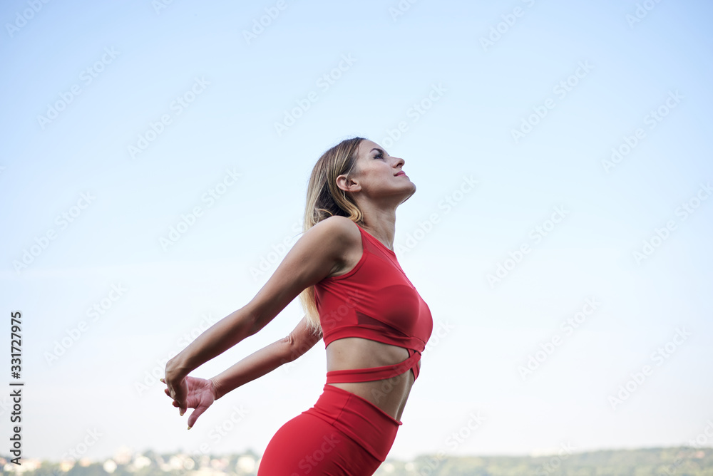Young blond woman, wearing red leggings, top and white sneakers, doing morning exercises by lake in summer. Fitness yoga stretching training outside by water. Healthy life concept. Active way of life Young blond woman, wearing red leggings, top and white sneakers, doing morning exercises by lake in summer. Fitness yoga stretching training outside by water. Healthy life concept. Active way of life