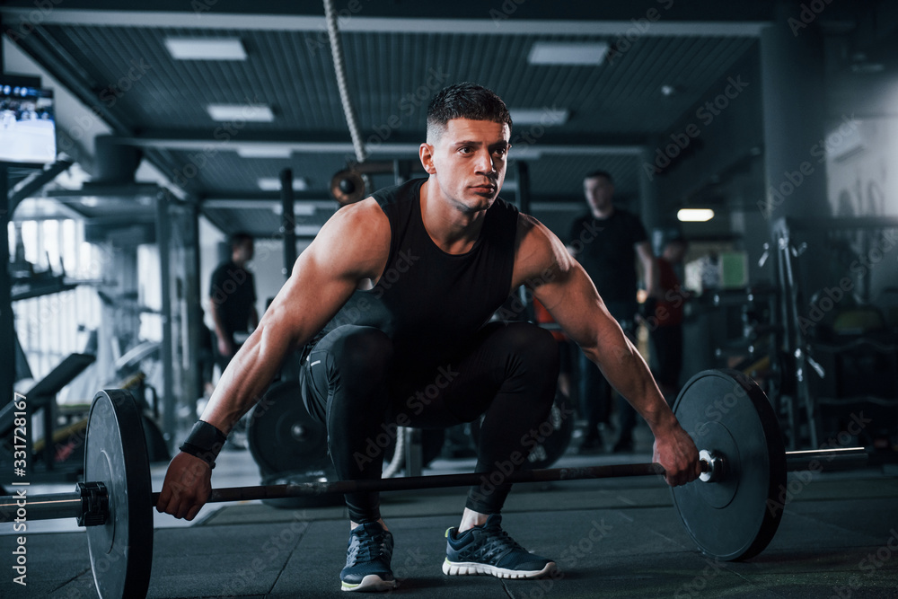 Strong young man in sportive clothes doing exercises with heavy weights in the gym