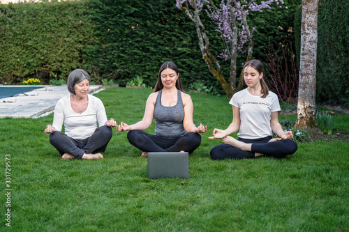 Mother, grandmother and teen daughter practicing online yoga class outdoors in garden at quarantine isolation period during coronavirus pandemic. Family doing sport together online from home. 