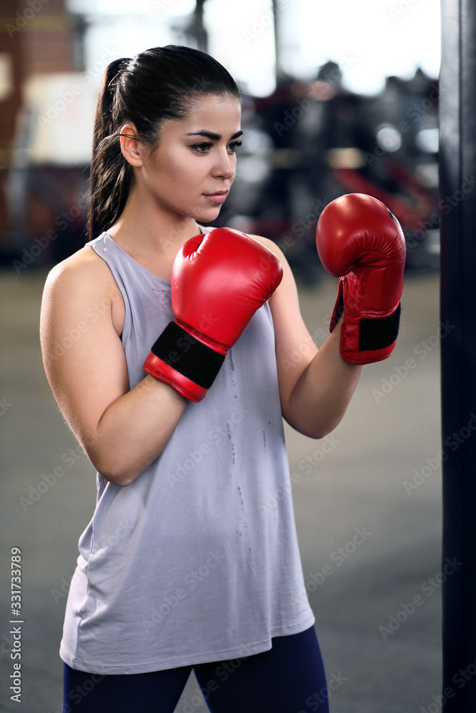 Athletic young brunette woman in sportswear and red boxing gloves trains bumps on a punching bag in a fitness gym.
