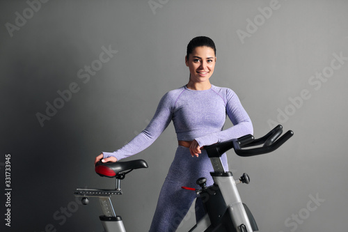 A beautiful athletic young brunette woman in sportswear trains on a sycle in the gym against the gray wall.