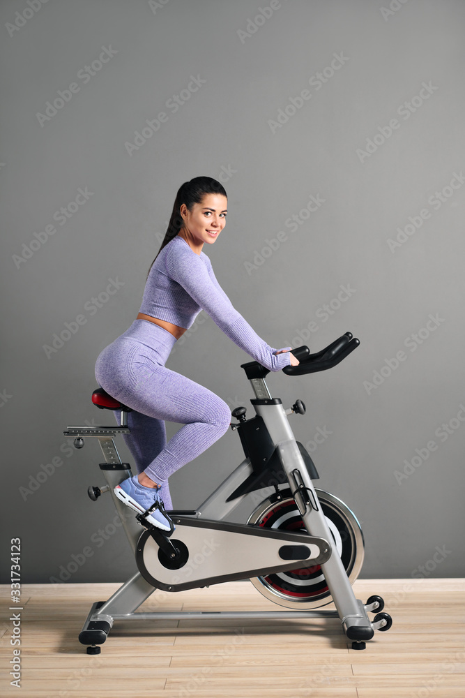 A beautiful athletic young brunette woman in sportswear trains on a sycle in the gym against the gray wall.