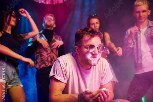 Young guy in sunglasses sniffing drug in night club with friends behind him