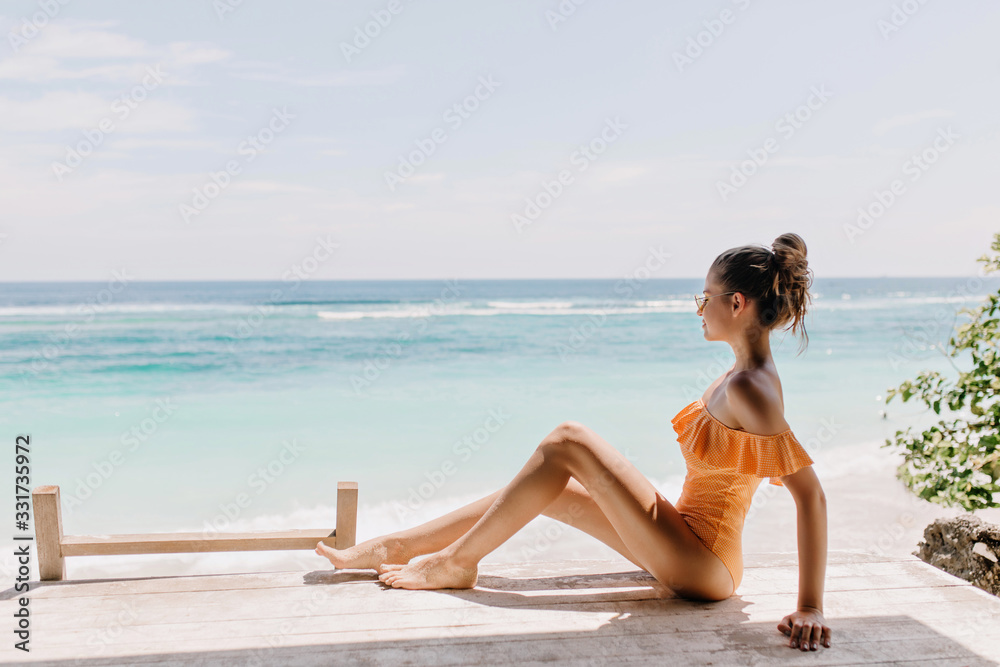 Pleasant woman in romantic swimsuit sitting on the ground and looking at horizon. Outdoor photo of slim white female model chilling at sea coast under clear sky. Pleasant woman in romantic swimsuit sitting on the ground and looking at horizon. Outdoor photo of slim white female model chilling at sea coast under clear sky.