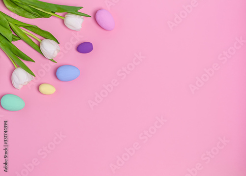 Colored Easter eggs and a bouquet of white tulips on a pink background, copy space, flat lay. Place for text. The concept of the holiday, spring. View from above.