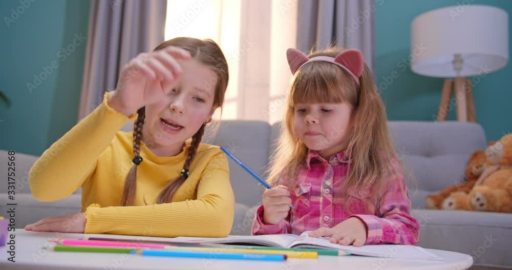 Joyful little Caucasian sisters staying at home alone and playing cheerfully together. Nice kids coloring picture with colorful pencils in cozy room. Happy children play and talking indoors. Joyful little Caucasian sisters staying at home alone and playing cheerfully together. Nice kids coloring picture with colorful pencils in cozy room. Happy children play and talking indoors.