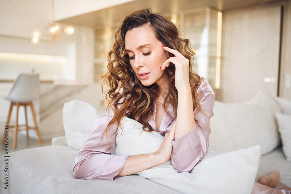 Beautiful young woman with curly hair in pink pajamas sitting at white couch or sofa in the morning. Scandinavian style living room & kitchen interior. Enjoying time at home. Furniture store concept Beautiful young woman with curly hair in pink pajamas sitting at white couch or sofa in the morning. Scandinavian style living room & kitchen interior. Enjoying time at home. Furniture store concept