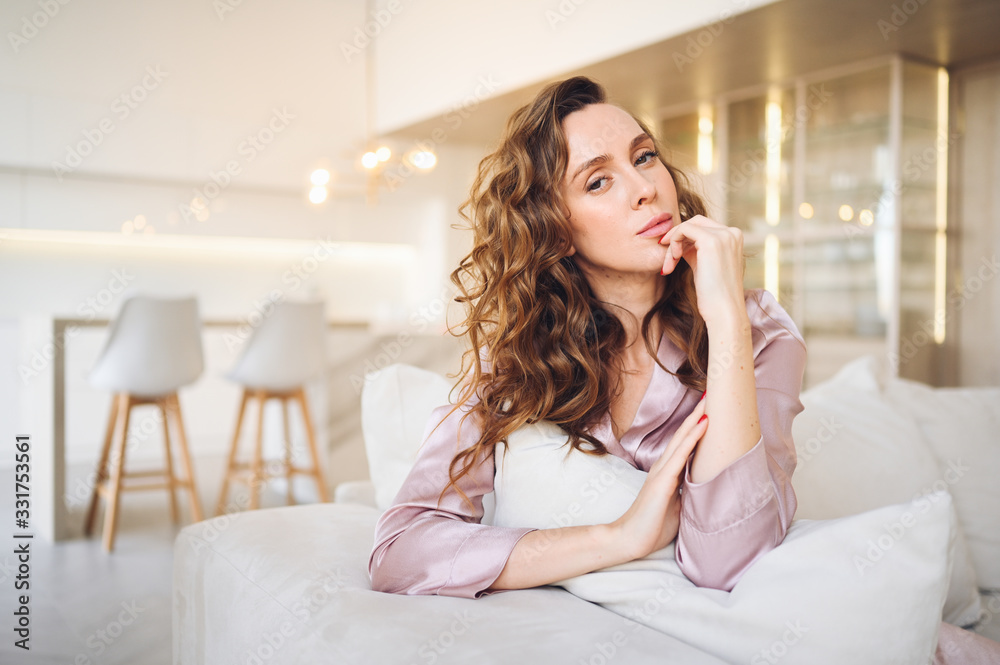 Beautiful young woman with curly hair in pink pajamas sitting at white couch or sofa in the morning. Scandinavian style living room & kitchen interior. Enjoying time at home. Furniture store concept Beautiful young woman with curly hair in pink pajamas sitting at white couch or sofa in the morning. Scandinavian style living room & kitchen interior. Enjoying time at home. Furniture store concept