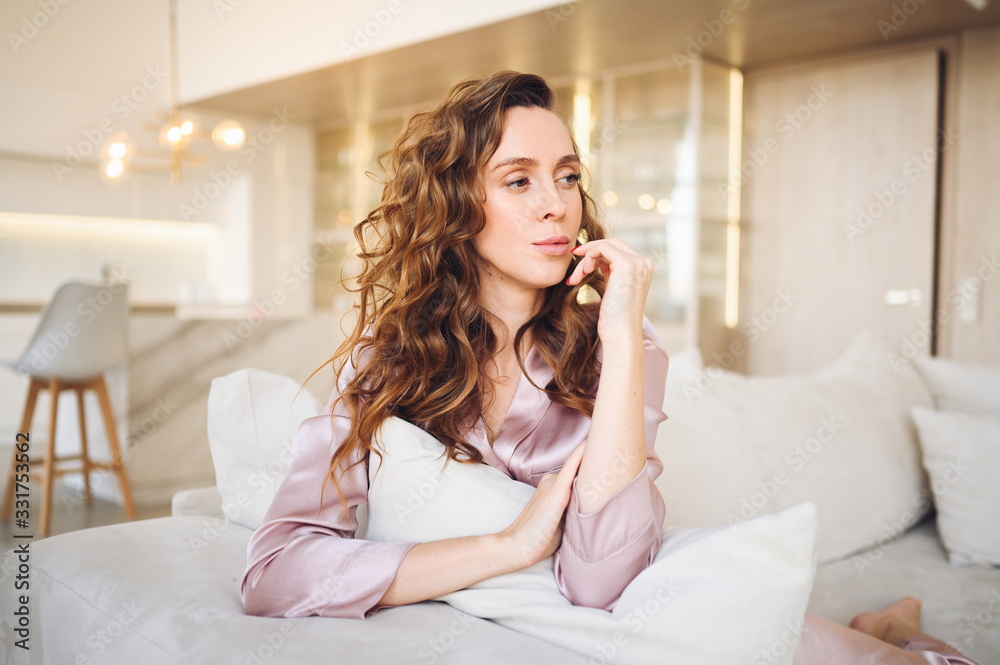 Beautiful young woman with curly hair in pink pajamas sitting at white couch or sofa in the morning. Scandinavian style living room & kitchen interior. Enjoying time at home. Furniture store concept Beautiful young woman with curly hair in pink pajamas sitting at white couch or sofa in the morning. Scandinavian style living room & kitchen interior. Enjoying time at home. Furniture store concept