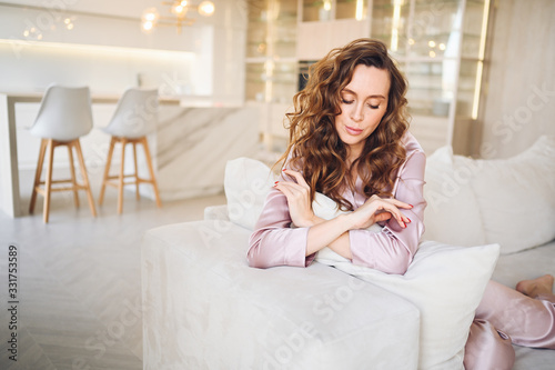 Beautiful young woman with curly hair in pink pajamas sitting at white couch or sofa in the morning. Scandinavian style living room & kitchen interior. Enjoying time at home. Furniture store concept