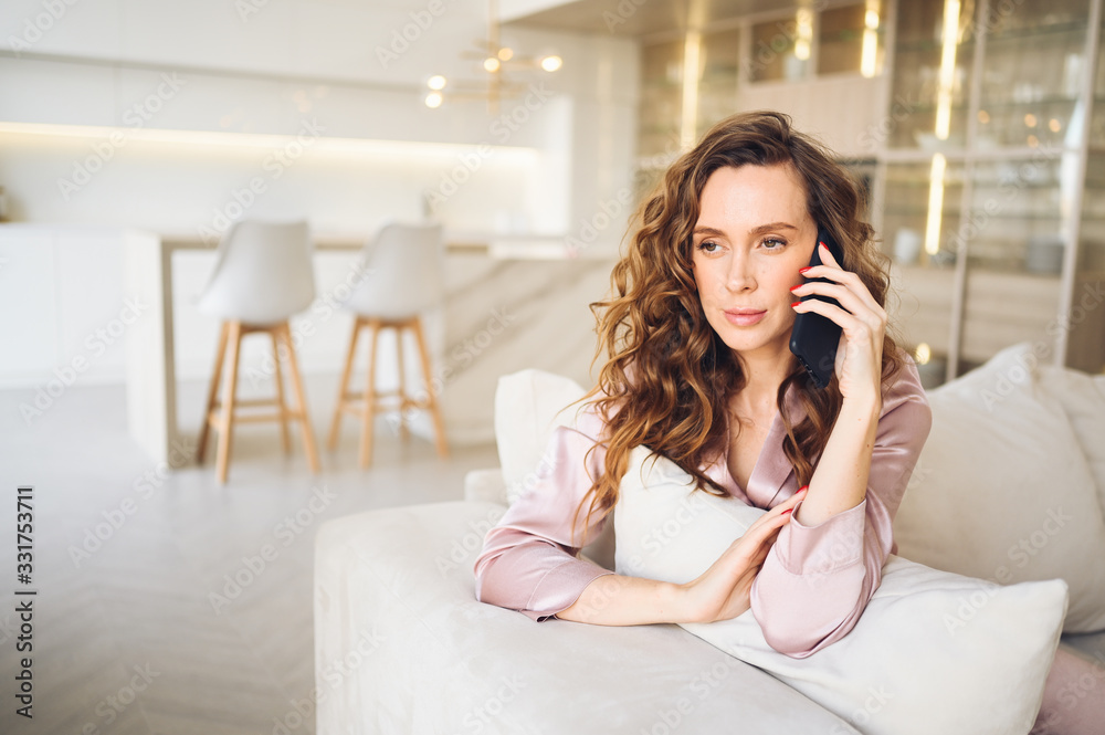 Beautiful young woman with curly hair in pink pajamas at white couch in the morning. Lady speaking phone in scandinavian style living room interior. Distance work from home. Quarantine COVID-19 Beautiful young woman with curly hair in pink pajamas at white couch in the morning. Lady speaking phone in scandinavian style living room interior. Distance work from home. Quarantine COVID-19