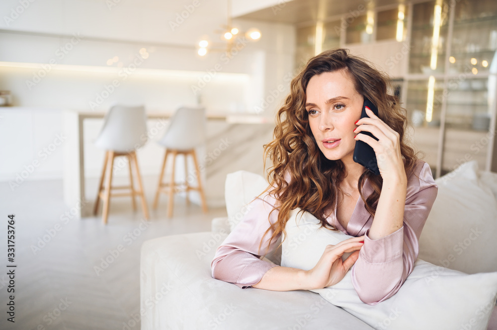 Beautiful young woman with curly hair in pink pajamas at white couch in the morning. Lady speaking phone in scandinavian style living room interior. Distance work from home. Quarantine COVID-19 Beautiful young woman with curly hair in pink pajamas at white couch in the morning. Lady speaking phone in scandinavian style living room interior. Distance work from home. Quarantine COVID-19