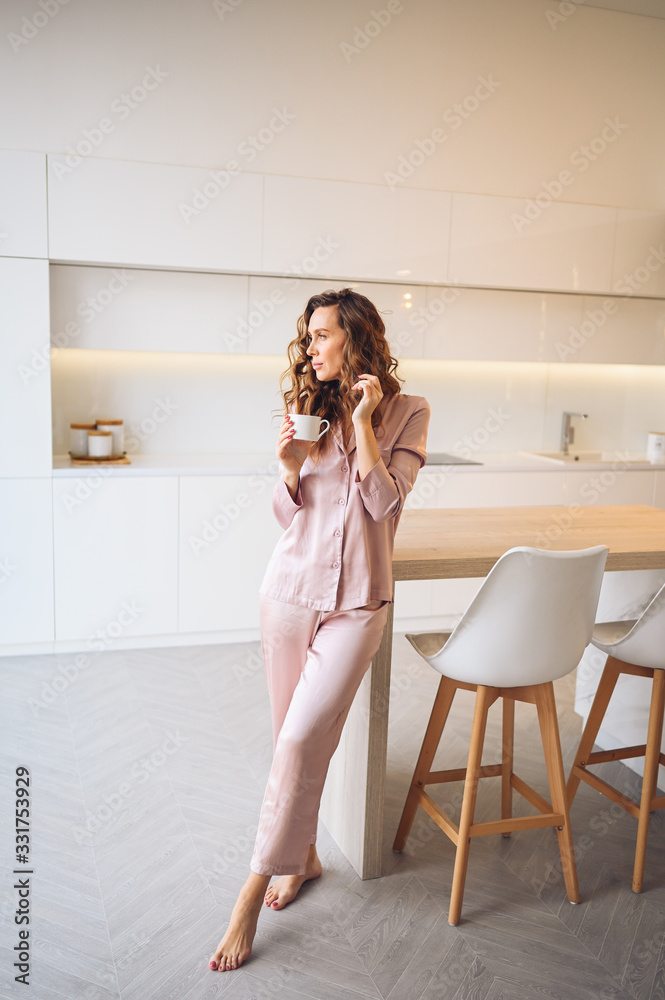 Beautiful young woman with curly hair drinking coffee in weekend morning in cozy pink pajamas. Lady in scandinavian style white kitchen modern interior. Home quarantine COVID-19 coronavirus pandemic Beautiful young woman with curly hair drinking coffee in weekend morning in cozy pink pajamas. Lady in scandinavian style white kitchen modern interior. Home quarantine COVID-19 coronavirus pandemic