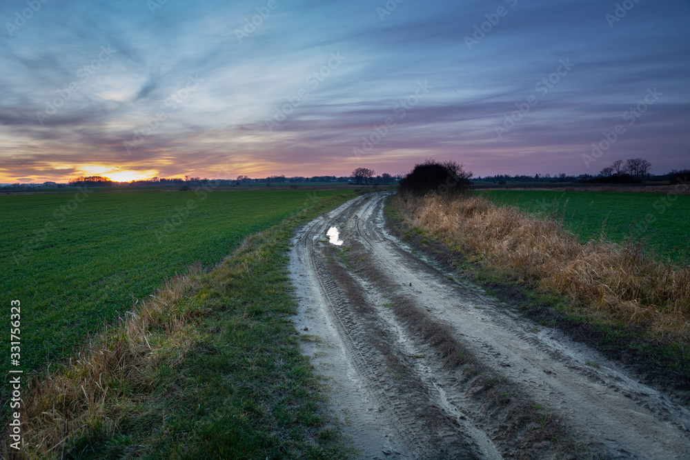 Dirt road through fields and clouds on sky after sunset