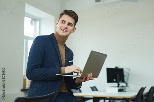 Young smiling businessman standing and holding laptop computer in his hands he working online at office