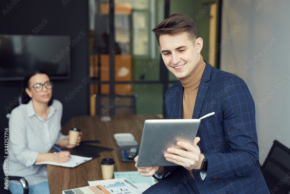 Young businessman holding tablet pc and using it for online communication with businesswoman sitting in the background at office Young businessman holding tablet pc and using it for online communication with businesswoman sitting in the background at office