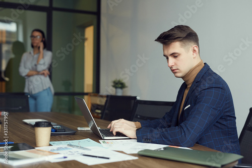 Young businessman sitting at the table typing on laptop computer and working with documents with businesswoman standing in the background at office