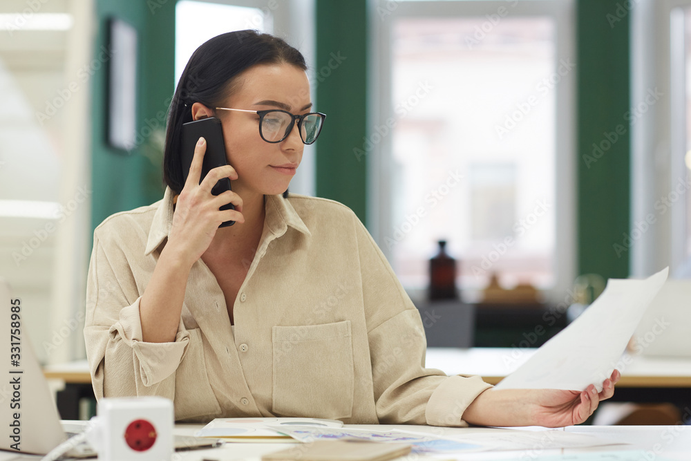 Young businesswoman in eyeglasses sitting at the table reading a document and talking on mobile phone at the office Young businesswoman in eyeglasses sitting at the table reading a document and talking on mobile phone at the office