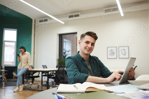 Portrait of young office worker smiling at camera while working with digital tablet at the table with his colleague in the background at office