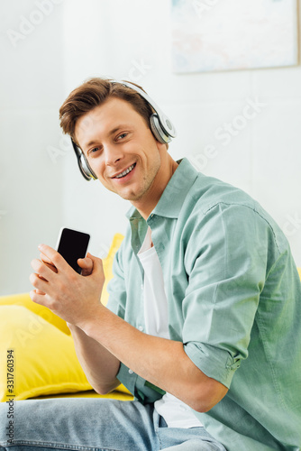 Side view of man in headphones smiling at camera and holding smartphone on couch