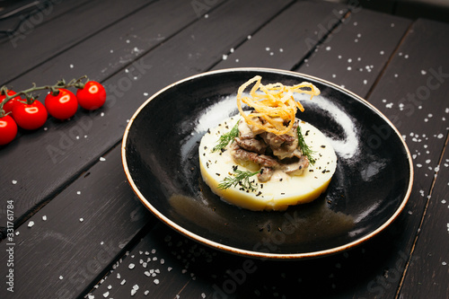Meat on a plate of tender mashed potatoes on a black glossy plate. The black table is sprinkled with salt and decorated with cherry tomatoes.