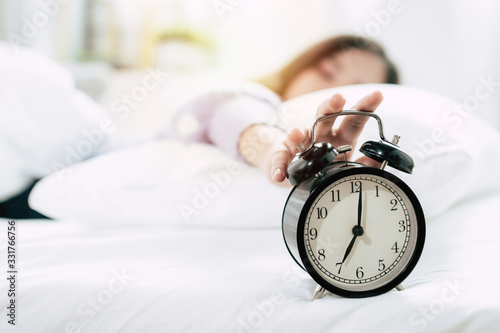 young asian woman sleeping on bed pressing snooze button on black vintage alarm clock at seven o'clock morning in bed room at home, lifestyle, good morning, healthy sleep and joyful weekend concept