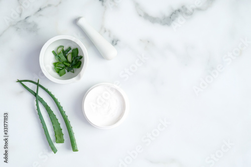 Cosmetic cream jar and green sliced stems aloe vera on marble background. Organic moisturizer face cream. Facial skin care and beauty treatment concept