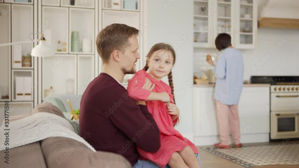 Little girl running to father sitting on sofa at home and coding on laptop computer. Side view of dad closing laptop, taking girl on lap and talking to her, mother cooking in kitchen in background