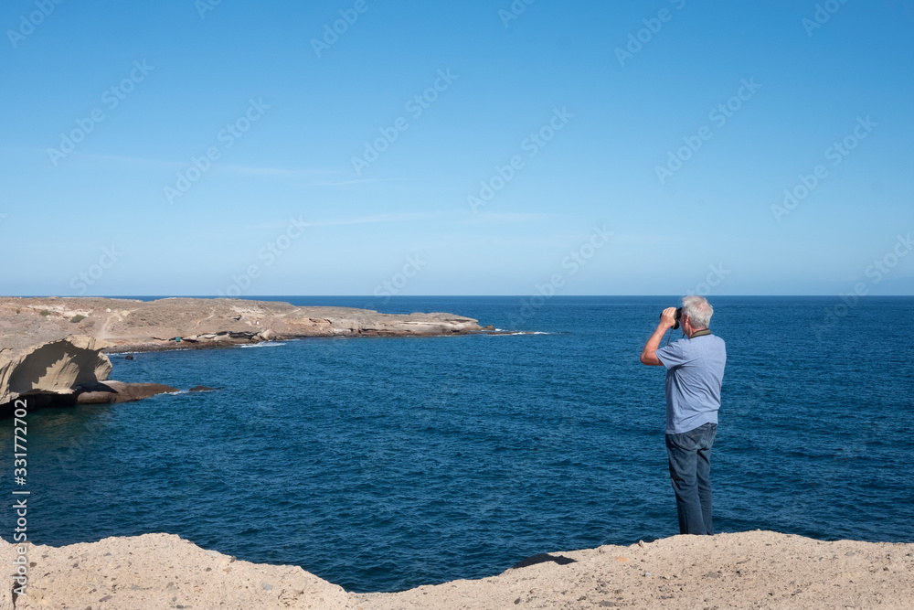 Rear view of elderly man looking with binoculars standing on the cliff - landscape of lava rocks and blue ocean - free and active retirement concept Rear view of elderly man looking with binoculars standing on the cliff - landscape of lava rocks and blue ocean - free and active retirement concept