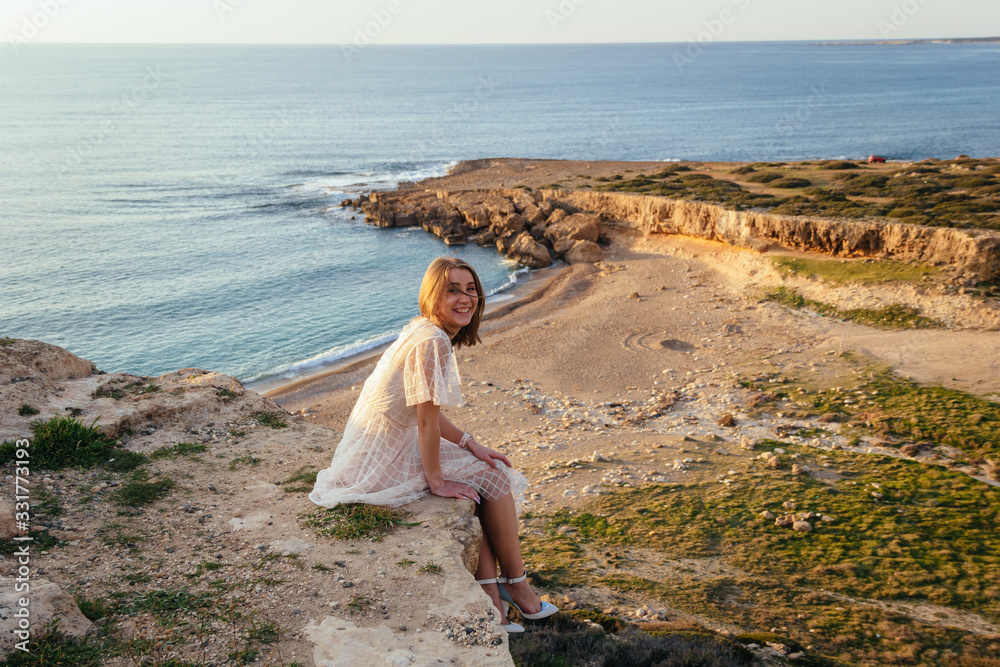 Gorgeous smiling woman in pink dress sits at the edge of cliff and feeling sea breeze at her smiling face. Sunset by the sea.