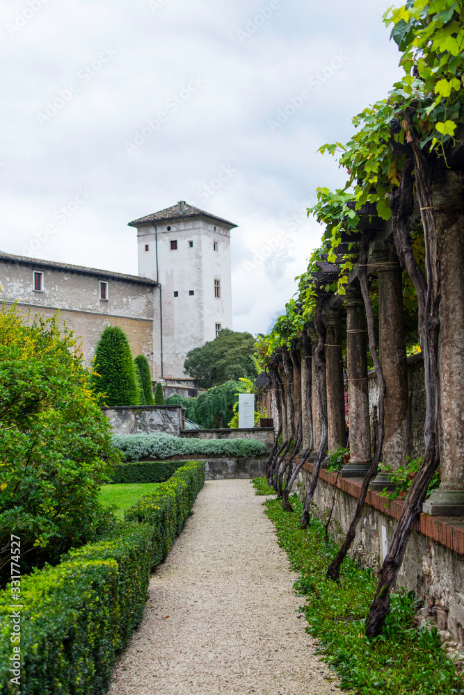 Inner courtyard of the historic castle complex in Trento, Italy. In the background is a tower to which the castle wall borders. On the left you can see a wooden pergola overgrown with wine.