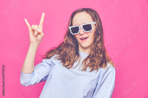 Stylish caucasian girl in a pale blue t-shirt and 8-bit glasses. A one-handed gesture is rock. Toned.