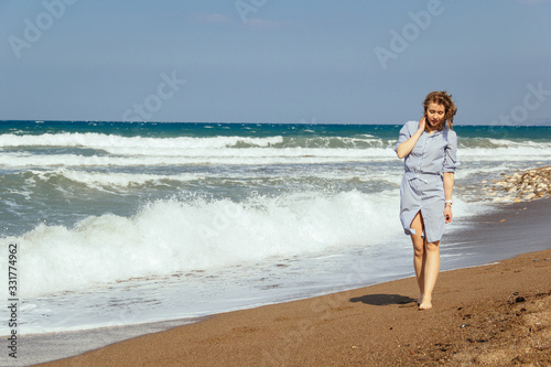 Gorgeous brown-haired girl in a striped dress walking at sand beach by the sea at sunny windy day