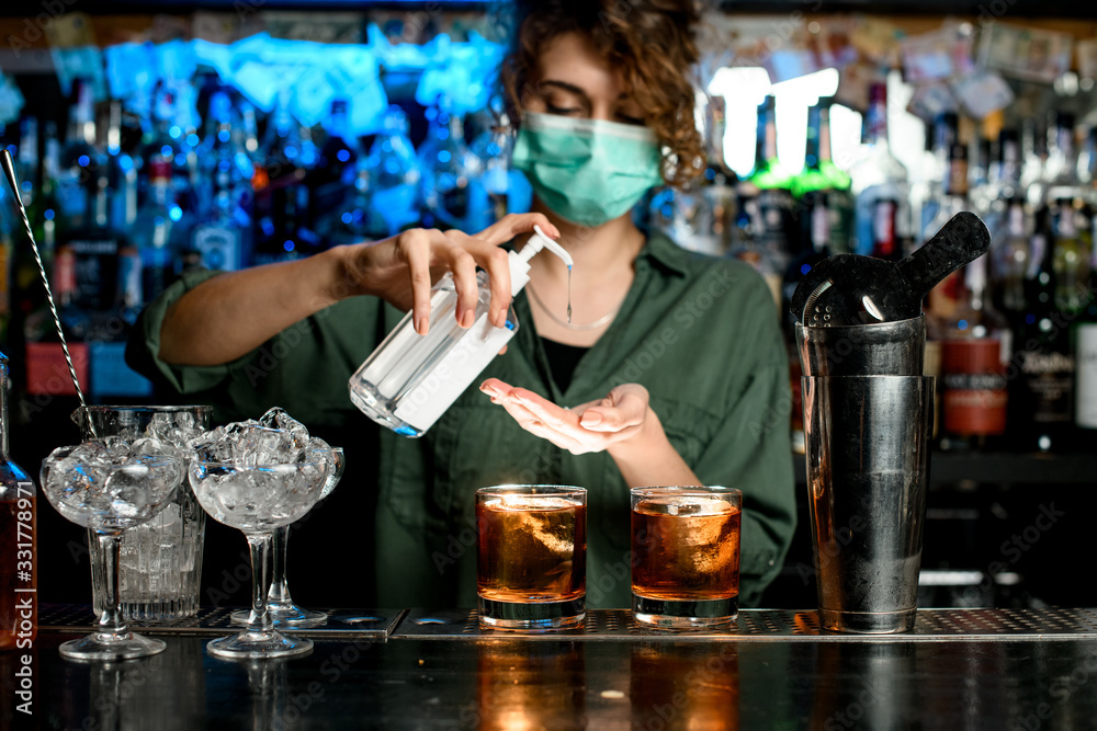 Woman bartender in medical mask treats her hands with disinfector - obrazy, fototapety, plakaty 