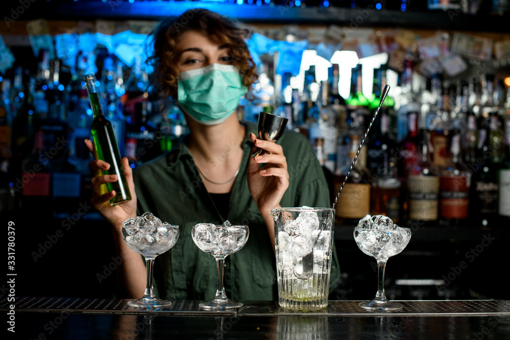 bartender girl in medical mask holding bottle of green liquid in one hand and beaker in other hand preparing to make mixture.
