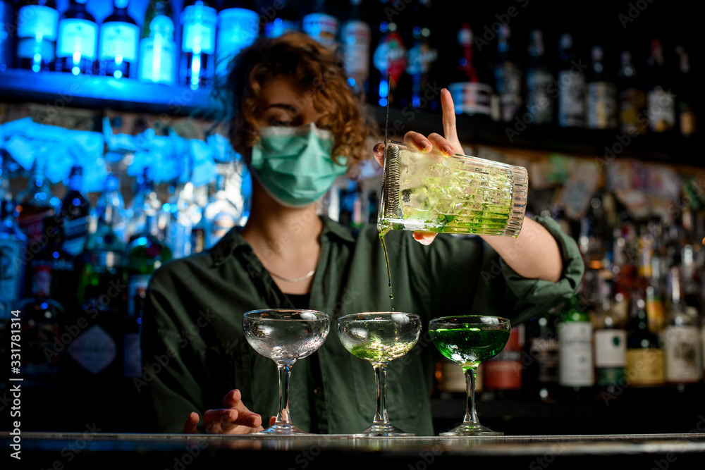 woman bartender in medical mask carefully pours drink from large glass into wineglasses.