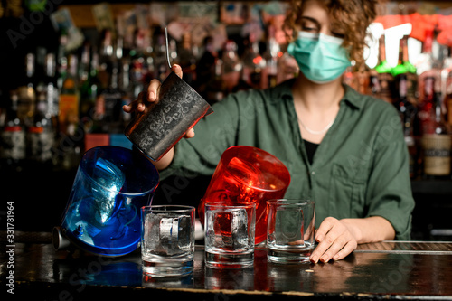 bartender girl in medical mask begin pour drink from steel glass into glasses with ice.