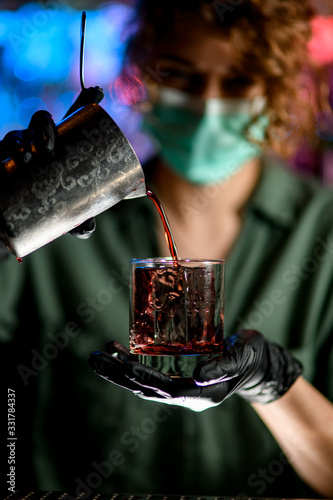 Close-up. Woman barman in medical mask and black gloves holds glass and attentively pours drink into it.