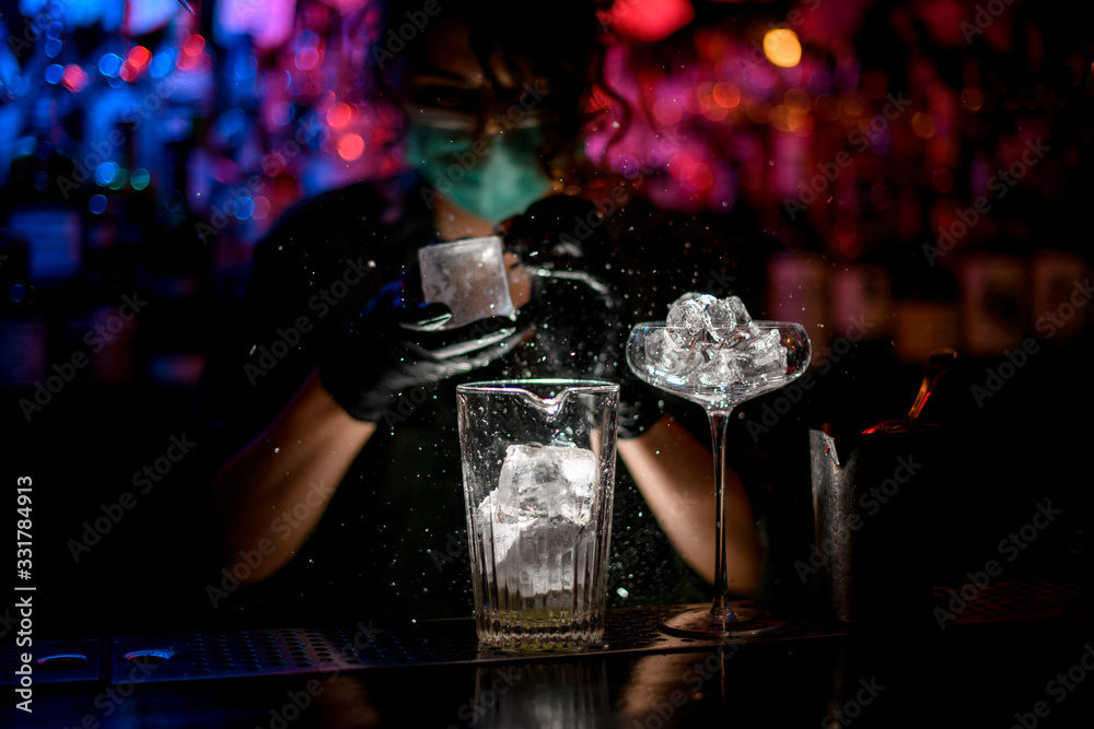 Close-up. Woman barman in medical mask and black gloves with special device breaks pieces of ice. - obrazy, fototapety, plakaty 