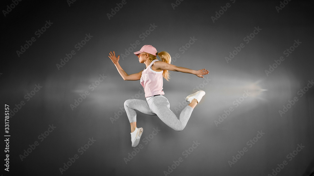 Young blonde woman with fit body jumping and running against grey background. Female model in sportswear exercising.