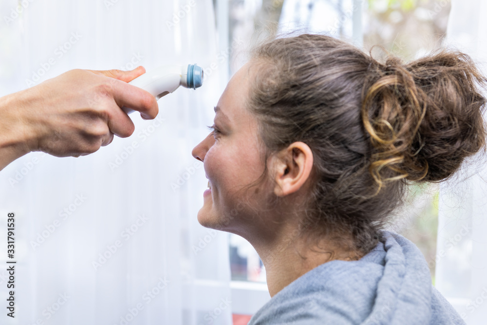 Doctor in white medical coat measuring temperature from young smiling woman using forehead digital thermometer in hospital room by window with bright natural light Doctor in white medical coat measuring temperature from young smiling woman using forehead digital thermometer in hospital room by window with bright natural light